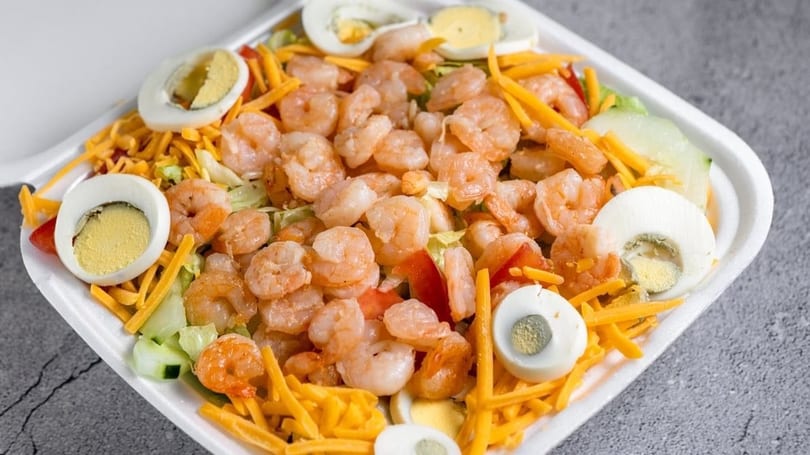 Shrimp salad with shredded cheese, hard-boiled eggs, and vegetables in a white takeout container.