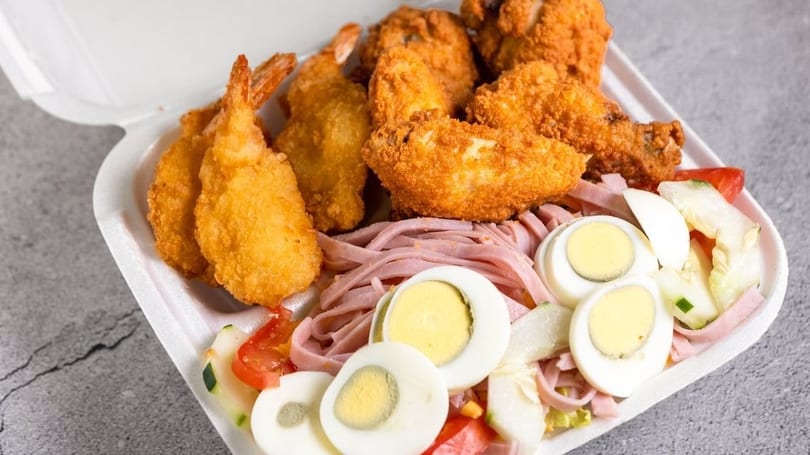A take-out container filled with fried chicken, fried shrimp, ham, sliced eggs, and vegetables.