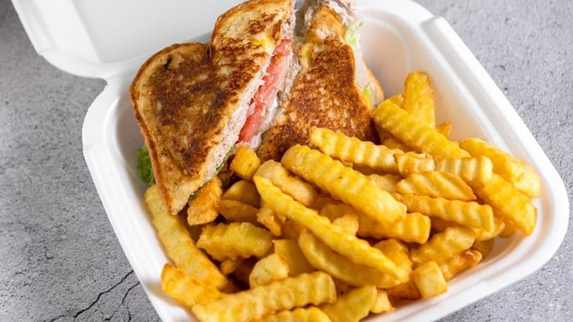 Grilled sandwich with tomatoes and lettuce, served with crinkle-cut fries in a white to-go container.