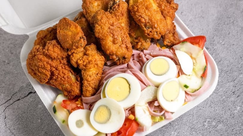 Fried chicken wings, sliced ham, hard-boiled eggs, and salad in a white takeout container.